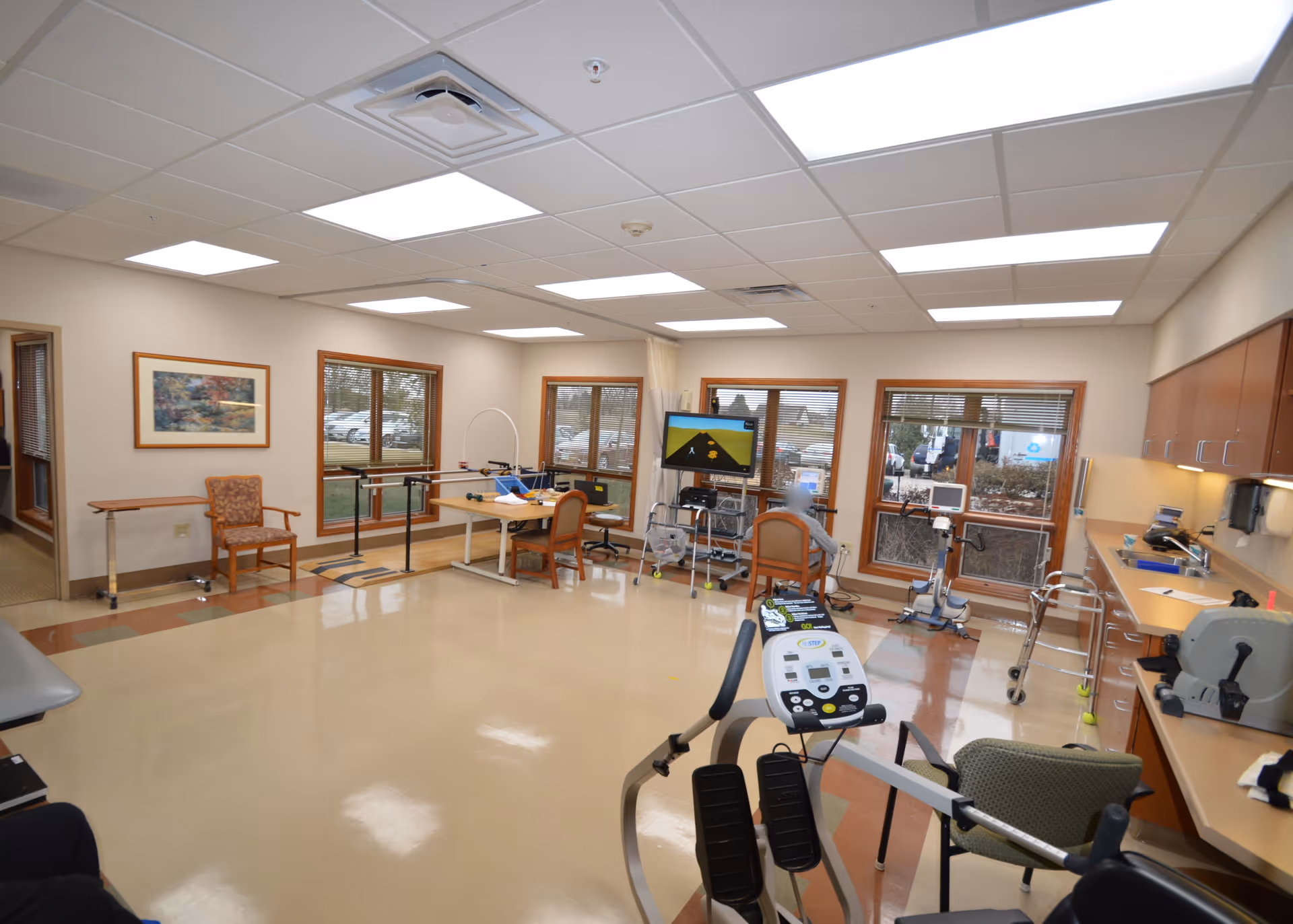 A spacious therapy or rehabilitation room with exercise equipment, chairs, tables, and a large screen displaying a game or exercise program. The room has large windows with a view outside, beige walls, and a tiled floor. There are cabinets and countertops along one wall with various supplies.
