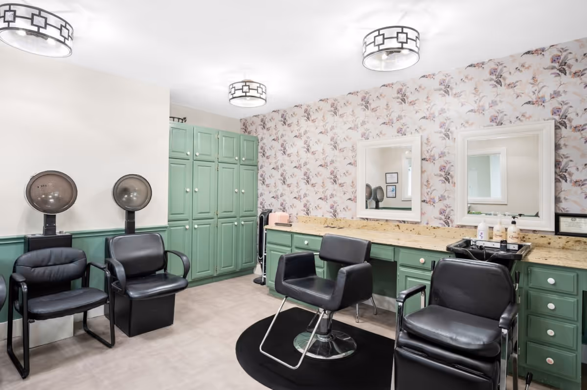 Interior view of a hair salon area in a senior living facility with two black salon chairs, two hair dryers, green cabinets, floral wallpaper, two mirrors, and ceiling lights.