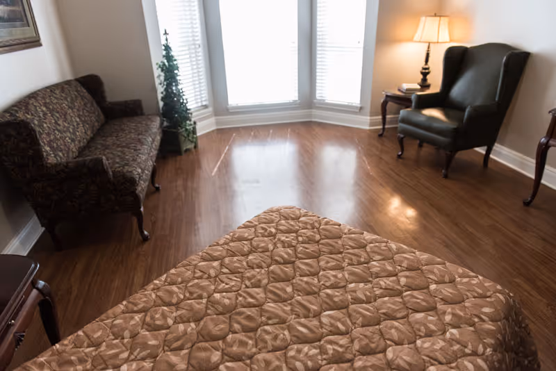 A cozy room with a patterned brown bedspread in the foreground, a floral-patterned sofa on the left, a green armchair with a side table and lamp on the right, and large windows with blinds letting in natural light.