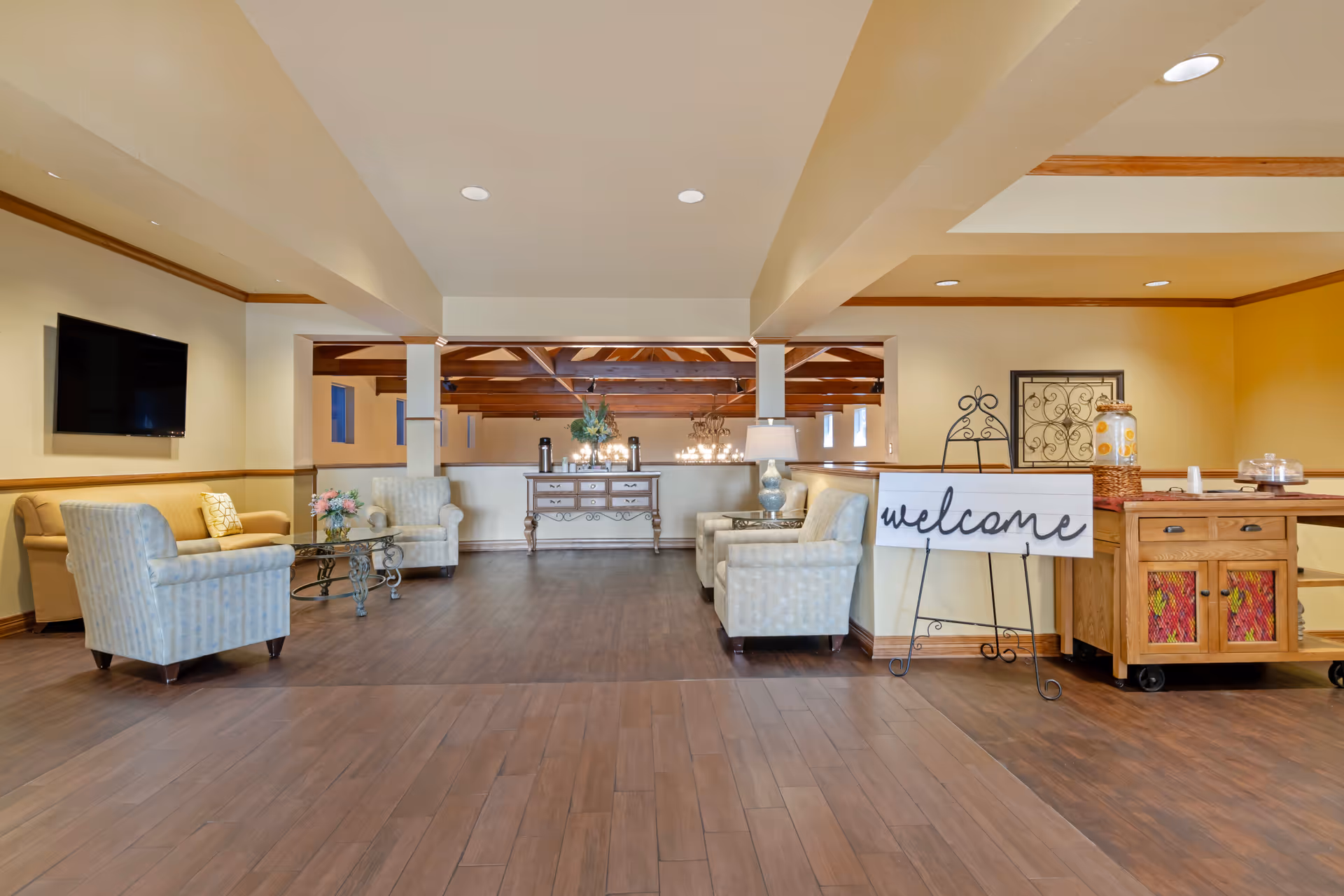 A spacious and well-lit common area with wooden flooring, beige walls, and recessed ceiling lights. The room features comfortable seating including two armchairs and a sofa arranged around a glass coffee table. A flat-screen TV is mounted on the wall to the left. In the background, there is a wooden console table with decorative items and a chandelier hanging from the ceiling. To the right, a wooden cart holds a beverage dispenser and a cake stand, next to a sign on an easel that reads 'welcome'.