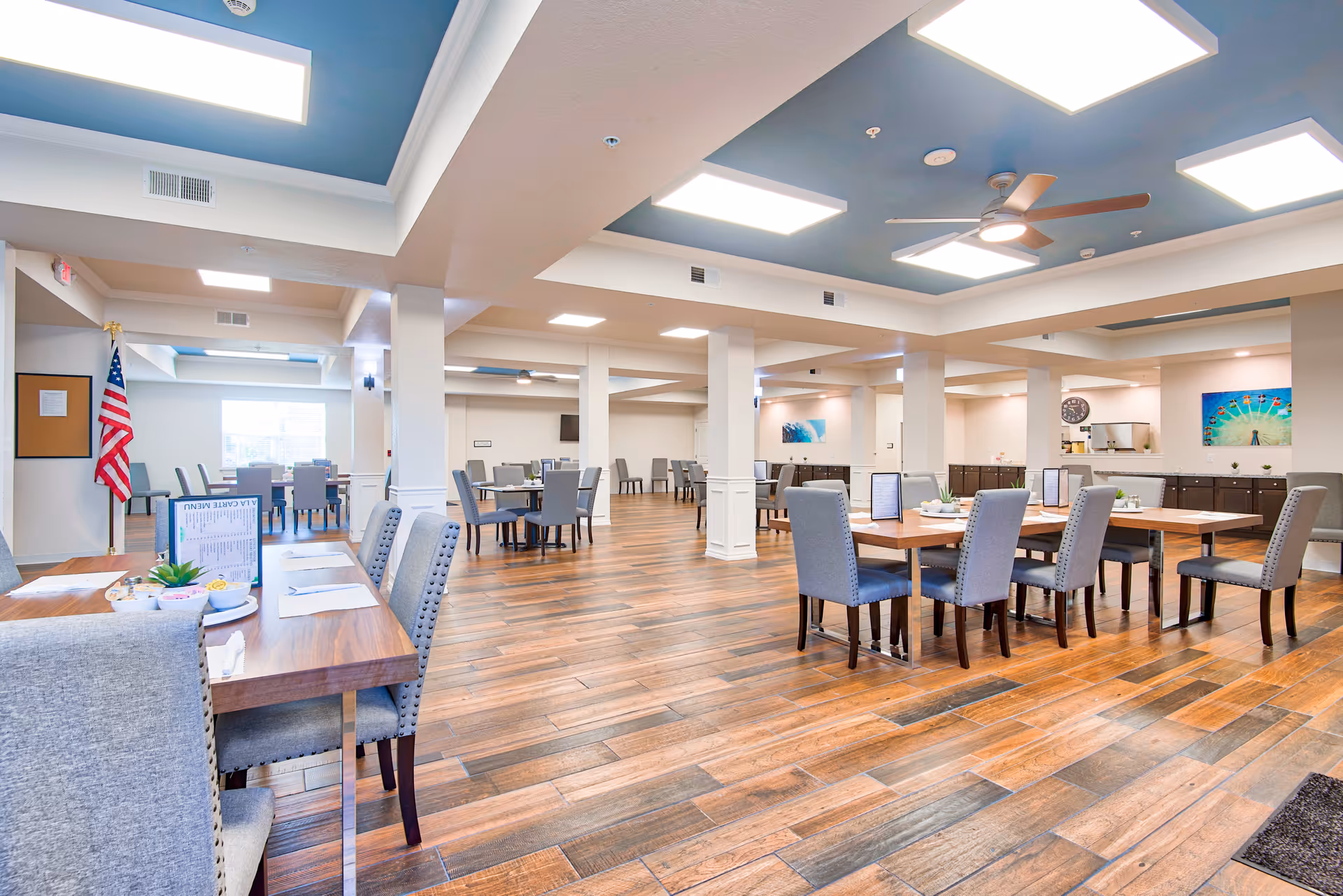 A spacious and well-lit dining room in The Carlstone Independent 55 Living facility featuring multiple wooden tables with gray upholstered chairs. The room has a wooden floor, white walls, and a ceiling with recessed lighting and ceiling fans. There are decorative paintings on the walls and an American flag near a window in the background.