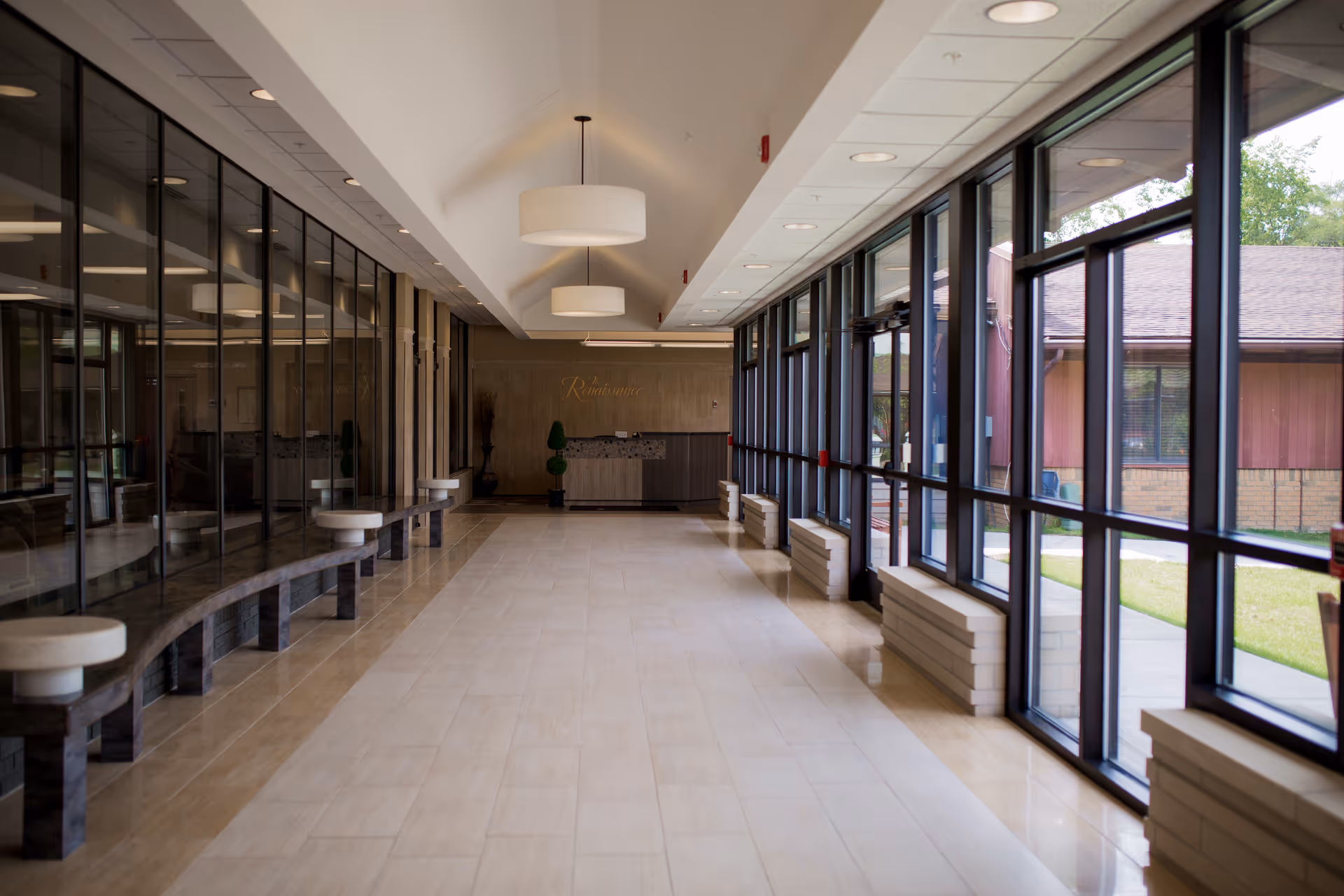A spacious, well-lit hallway with large floor-to-ceiling windows on the right side showing an outdoor view. On the left side, there is a long curved bench with round white cushions. The ceiling has recessed lighting and two large hanging light fixtures. At the end of the hallway, there is a reception desk with decorative plants and a sign that reads 'Renaissance'.