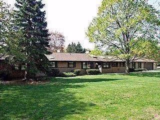 Single-story residential building surrounded by green grass and large trees under a clear sky.