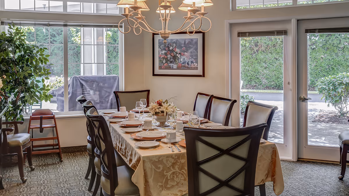 A dining room set for a meal with a long table covered with a beige tablecloth, plates, cups, glasses, and silverware. The room has large windows and glass doors letting in natural light, with greenery visible outside. There is a chandelier hanging above the table and a framed floral painting on the wall.