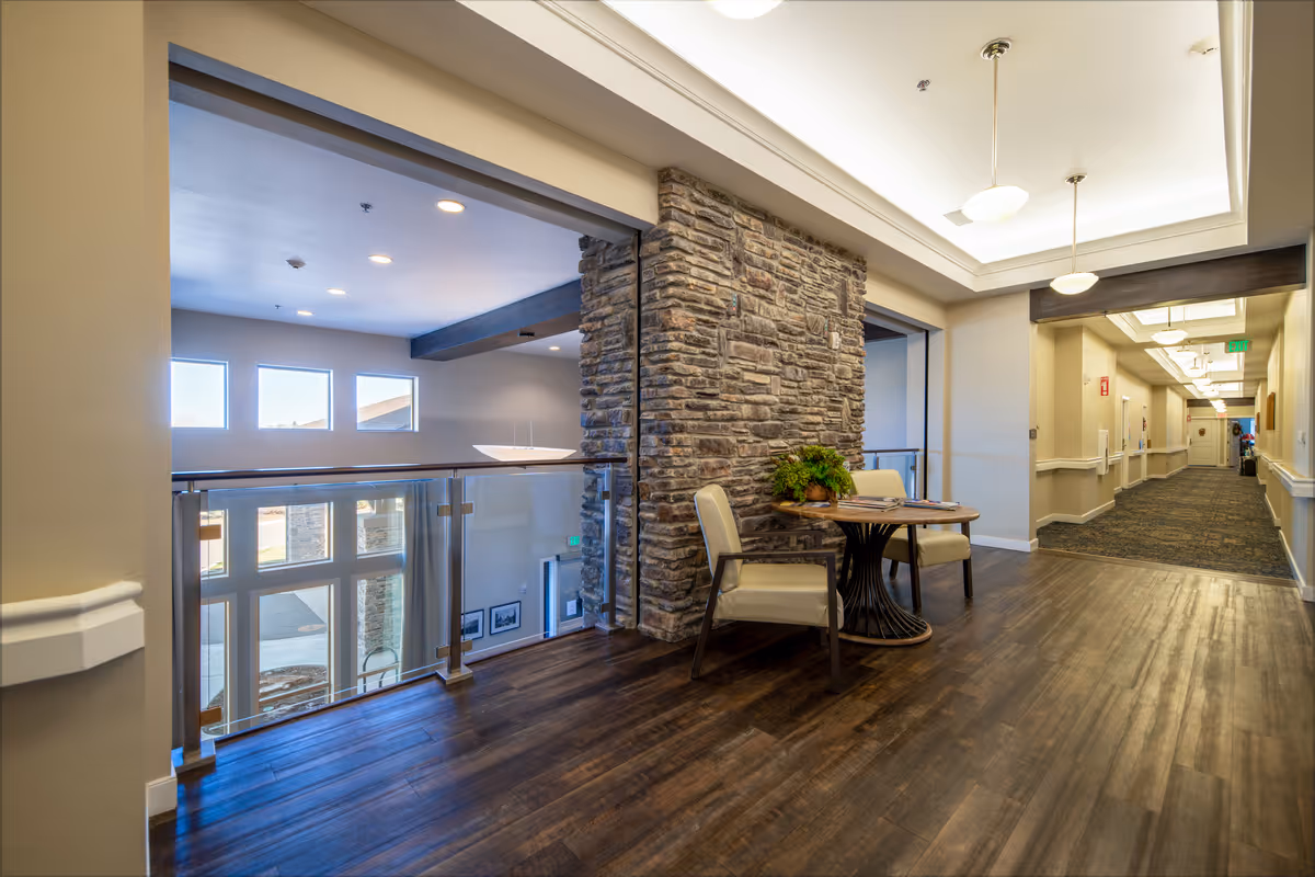Interior hallway area of a senior living facility with wood flooring, a stone accent wall, two chairs and a small round table with a plant and magazines. The hallway extends to the right with carpeted flooring and ceiling lights. Large windows provide natural light to the space.