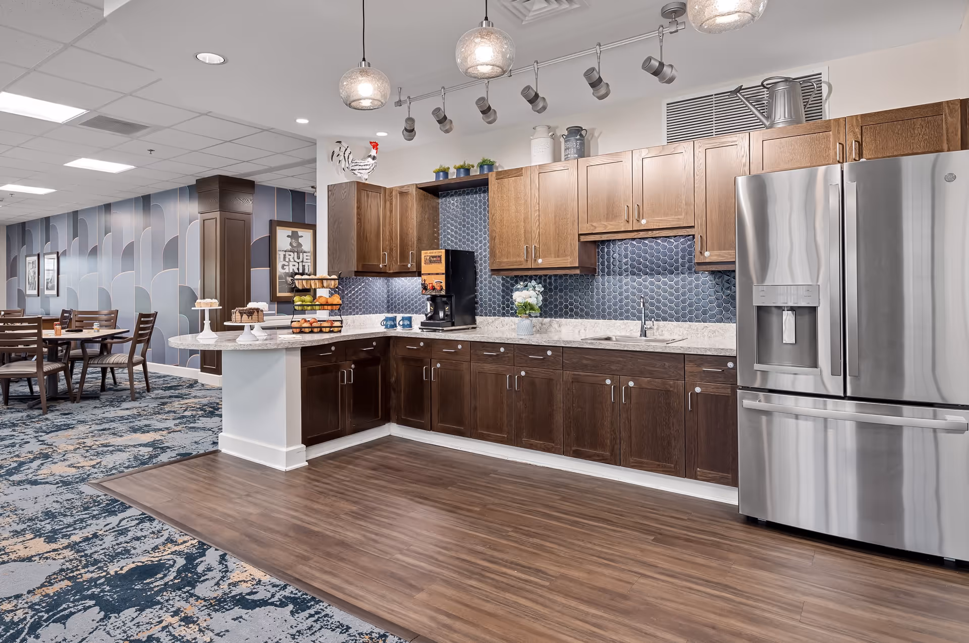 A modern kitchen area with wooden cabinets in two tones, a stainless steel refrigerator, a coffee maker on the counter, and a decorative backsplash with hexagonal tiles. The kitchen opens into a dining area with tables and chairs, and the floor transitions from wood to patterned carpet. The walls have a geometric wallpaper design, and there are pendant lights and track lighting above the kitchen counter.
