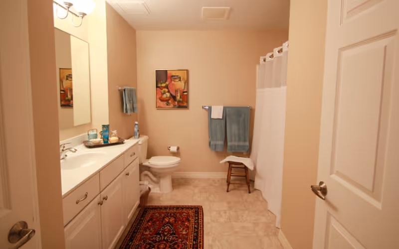 A clean bathroom with beige walls and tiled floor. There is a white vanity with a sink and mirror on the left side. A toilet is positioned in the center against the back wall, with a colorful framed painting above it. Two blue towels hang on a towel rack next to the toilet, and a small wooden stool with a white towel is placed near a shower with a white curtain on the right side. A decorative rug is on the floor in front of the vanity.