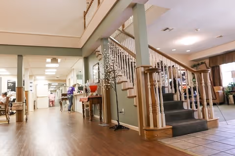 Interior view of a senior living facility hallway with wooden flooring, a staircase with wooden railings and carpeted steps, and a small table with a decorative tree and a drink dispenser. People are visible in the background near the hallway and seating area.