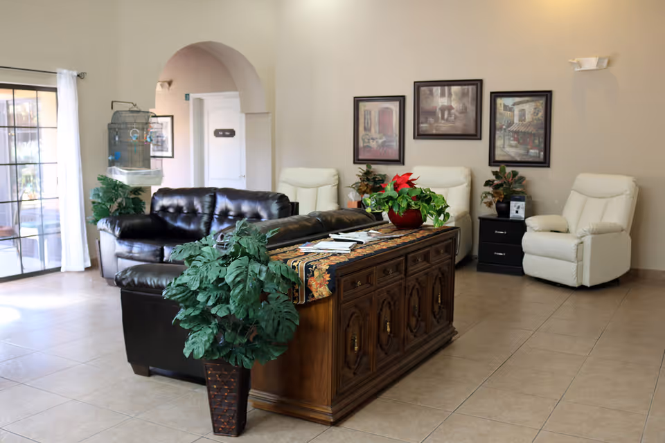 A cozy living room area in Carefree Assisted Living Center featuring a black leather sofa, two white recliner chairs, a wooden cabinet with decorative plants and magazines on top, framed artwork on the wall, and a large window with white curtains letting in natural light.