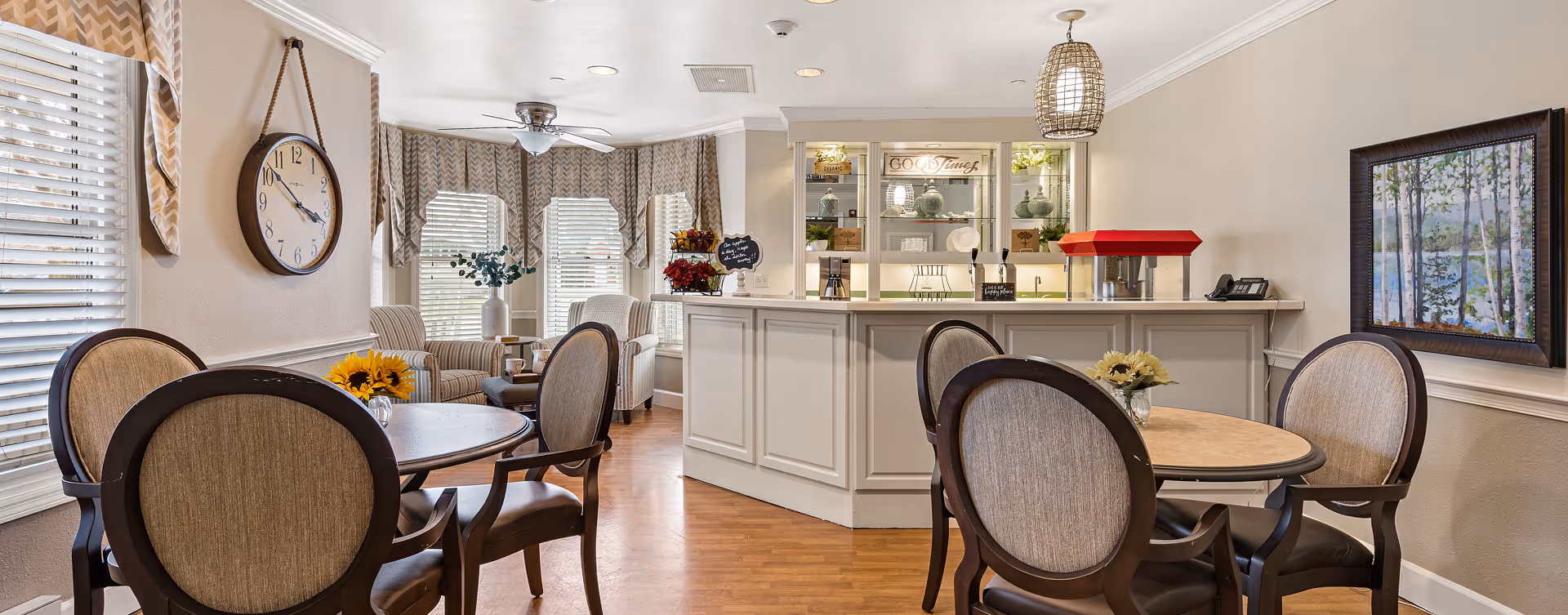 Bright common dining and seating area with round tables and upholstered chairs, a service counter and windows with valances.
