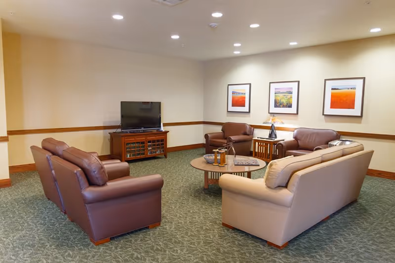 A cozy senior living facility lounge area with several leather armchairs and sofas arranged around a circular wooden coffee table. A flat-screen TV sits on a wooden cabinet against the wall. Three framed landscape paintings hang on the wall above a side table with a lamp. The room has beige walls, green patterned carpet, and recessed ceiling lights.