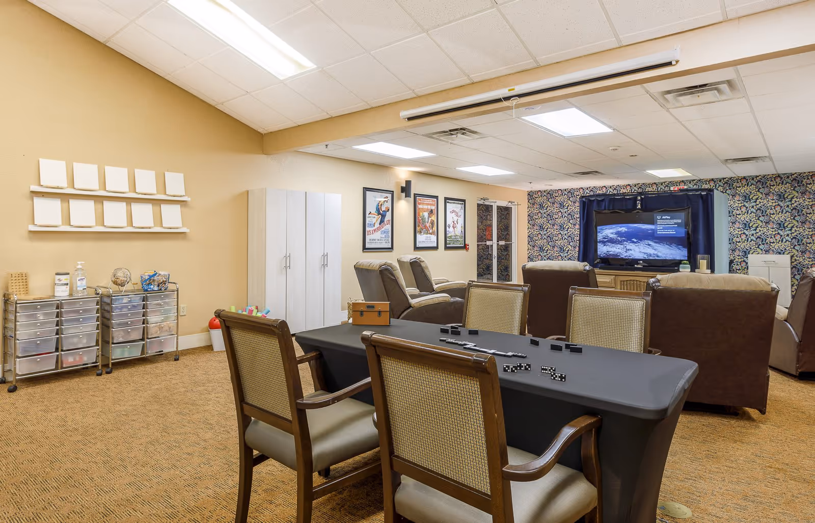 Community activity room with a table and chairs, recliners, and a large TV in a senior living facility.
