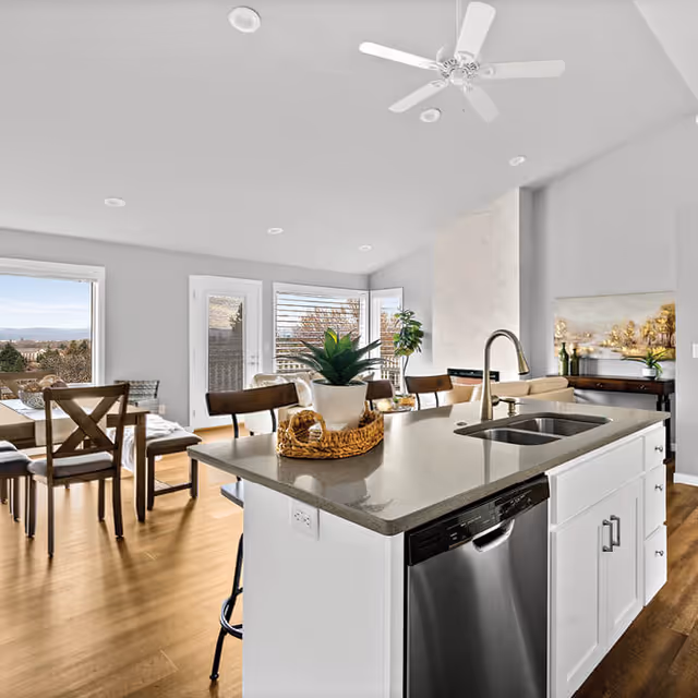 Bright and modern kitchen and dining area with a large island countertop featuring a sink and a potted plant centerpiece. The dining table has wooden chairs and is positioned near large windows and a glass door that provide a view of the outdoors. The room has light-colored walls, wooden flooring, recessed ceiling lights, and a ceiling fan.