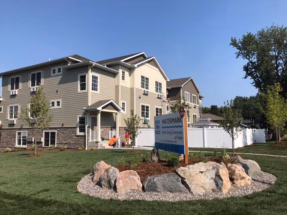 Exterior view of a two-story senior living community building with beige and brown siding, multiple windows, and a small porch entrance. In front of the building is a landscaped area with rocks and a sign that reads 'Watermark Senior Living Community of Fridley'. The sky is clear and blue, and there are a few trees and green grass surrounding the building.