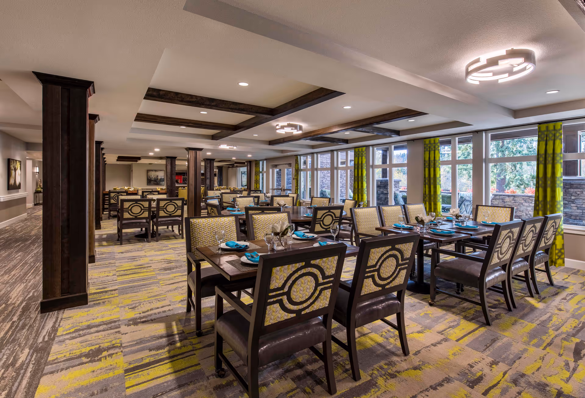 A spacious and well-lit dining room with multiple tables and chairs arranged neatly. Each table is set with blue napkins, glasses, and silverware. Large windows with green patterned curtains allow natural light to fill the room. The ceiling features recessed lighting and decorative wooden beams.