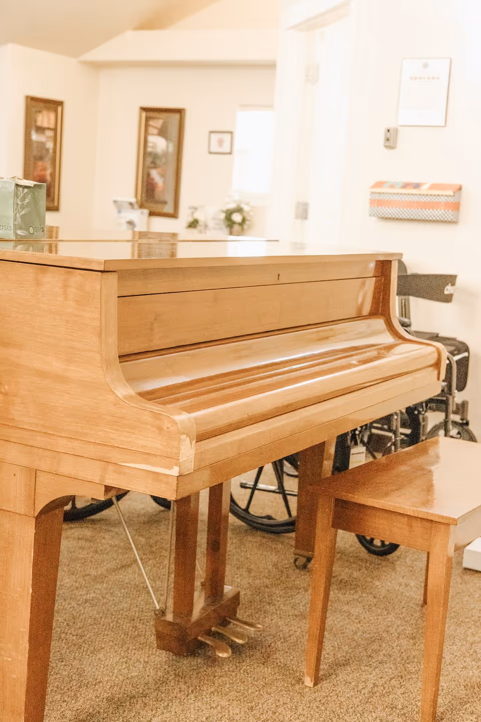 A wooden upright piano with a matching wooden bench in a carpeted room. In the background, there are framed pictures on the wall, a window, a wheelchair, and some decorative items on a table.