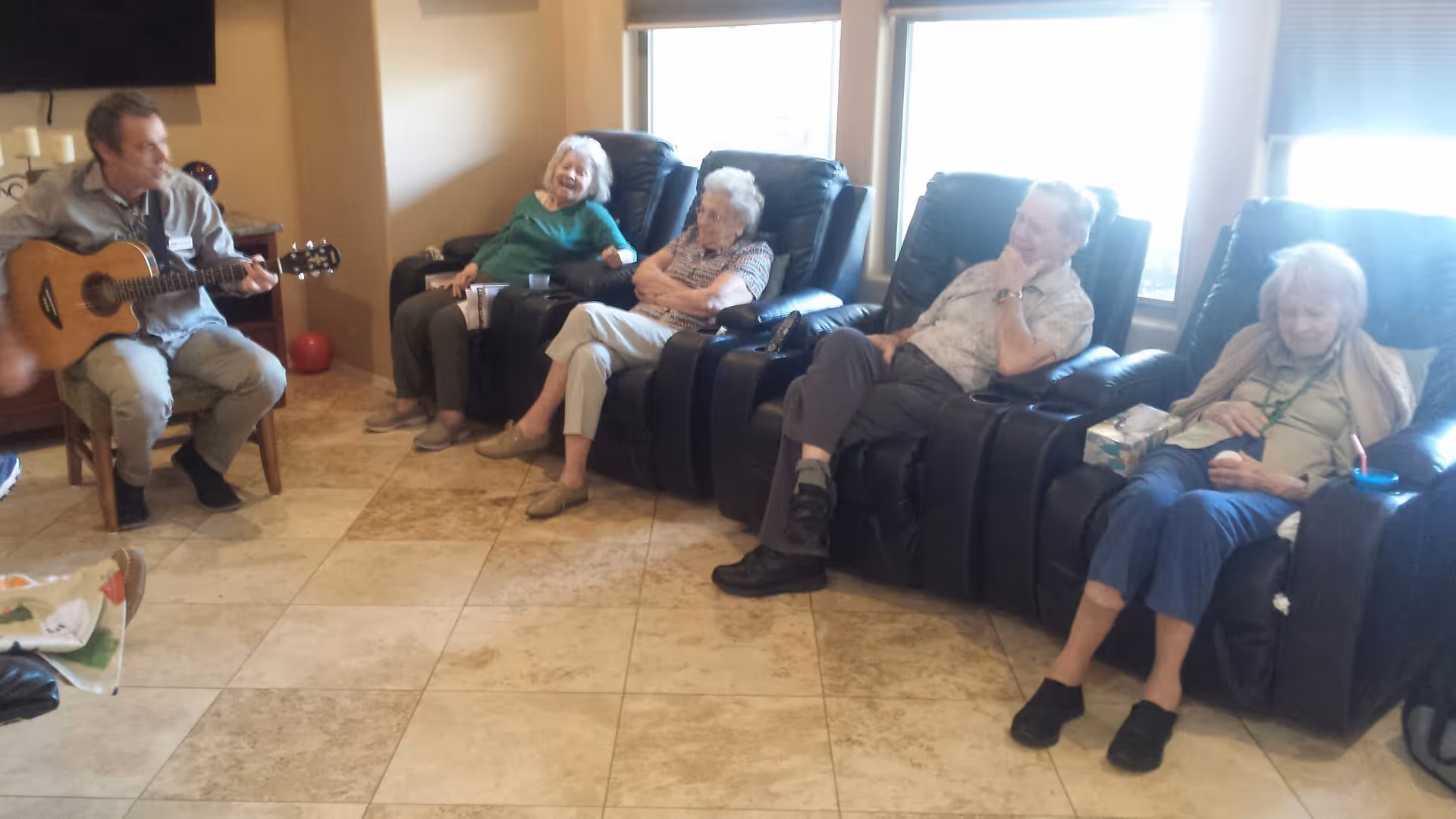 A group of elderly people sitting in black recliner chairs in a living room area, with a man playing guitar and engaging with them. The room has tiled flooring and large windows letting in natural light.