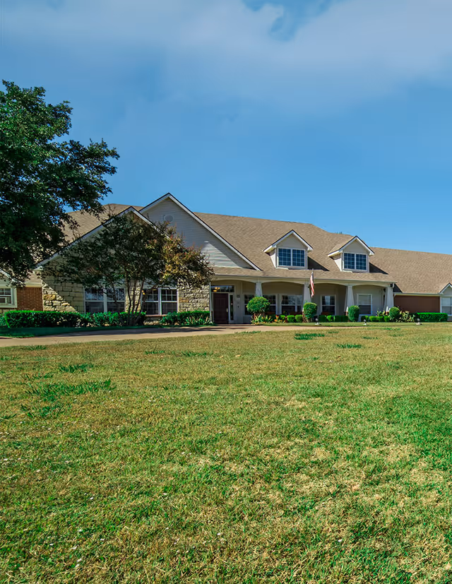 Exterior view of a single-story senior living facility building with a large front lawn, several windows, a covered entrance, and a few small trees and shrubs. The sky is clear and blue.