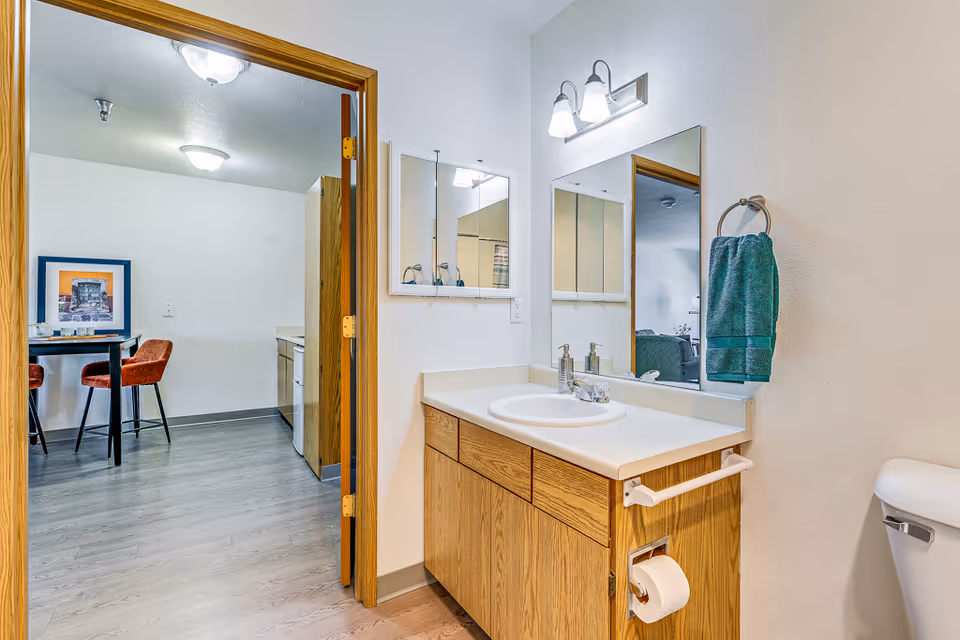 Bathroom with a wooden vanity featuring a white sink and countertop, a mirror above the sink, a green towel hanging on a ring, and a toilet partially visible. The bathroom door is open, showing a dining area with a table, two red chairs, and a framed picture on the wall.