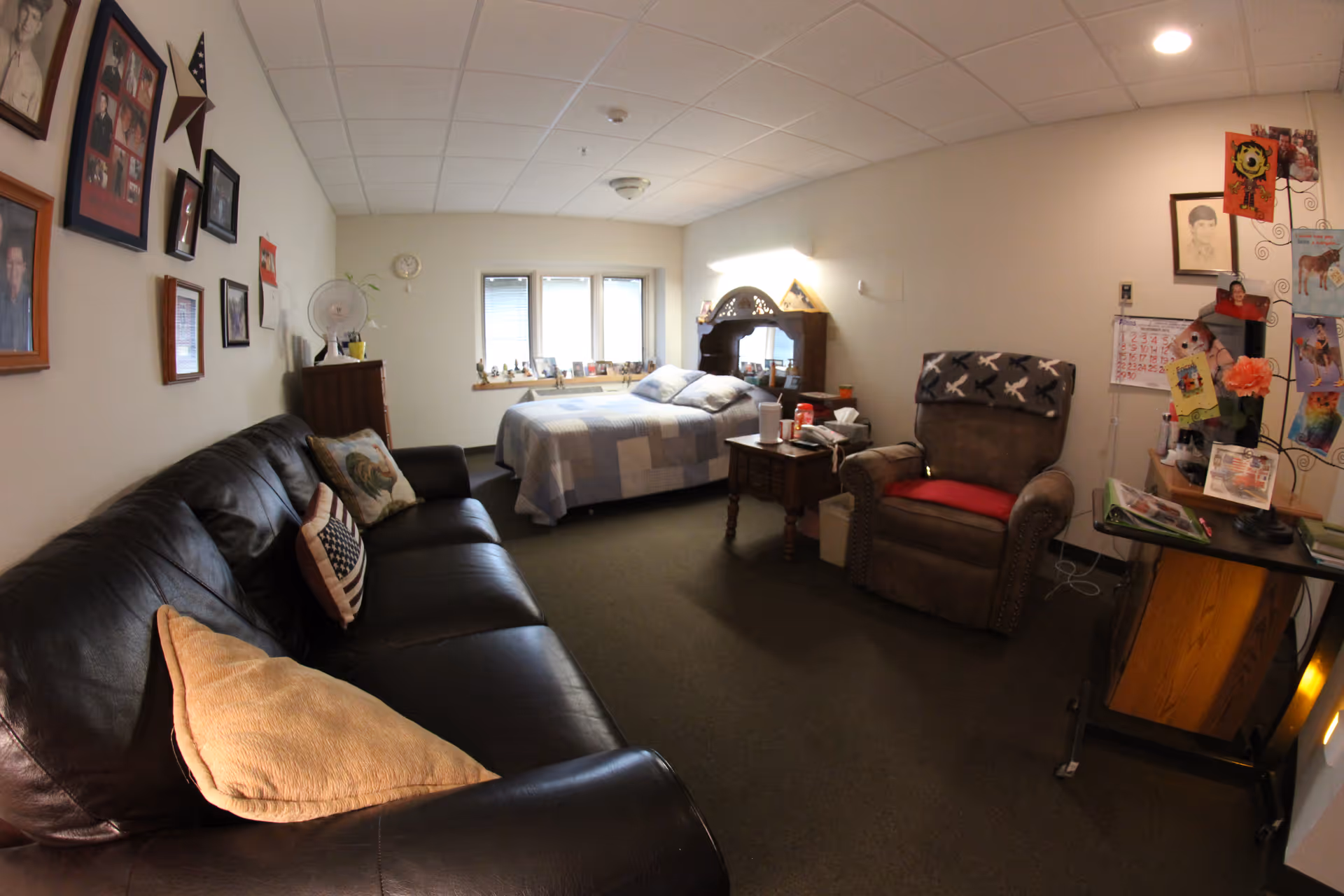 A cozy senior living room and bedroom combined space featuring a black leather couch with pillows, a comfortable armchair with a red cushion and a blanket draped over the back, a wooden side table with various items, and a bed with a patchwork quilt near a window. The walls are decorated with framed photos and artwork, and there is a small desk with personal items and decorations.