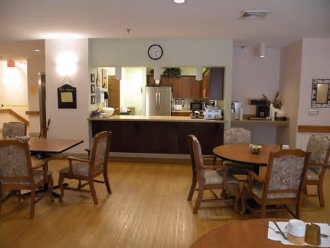 A dining area in a senior living facility with wooden tables and upholstered chairs. The room has a light wood floor and a counter window opening into a kitchen area with visible appliances including a refrigerator and microwave. A clock is mounted on the wall above the counter, and there are decorative items and a small chalkboard on the walls.