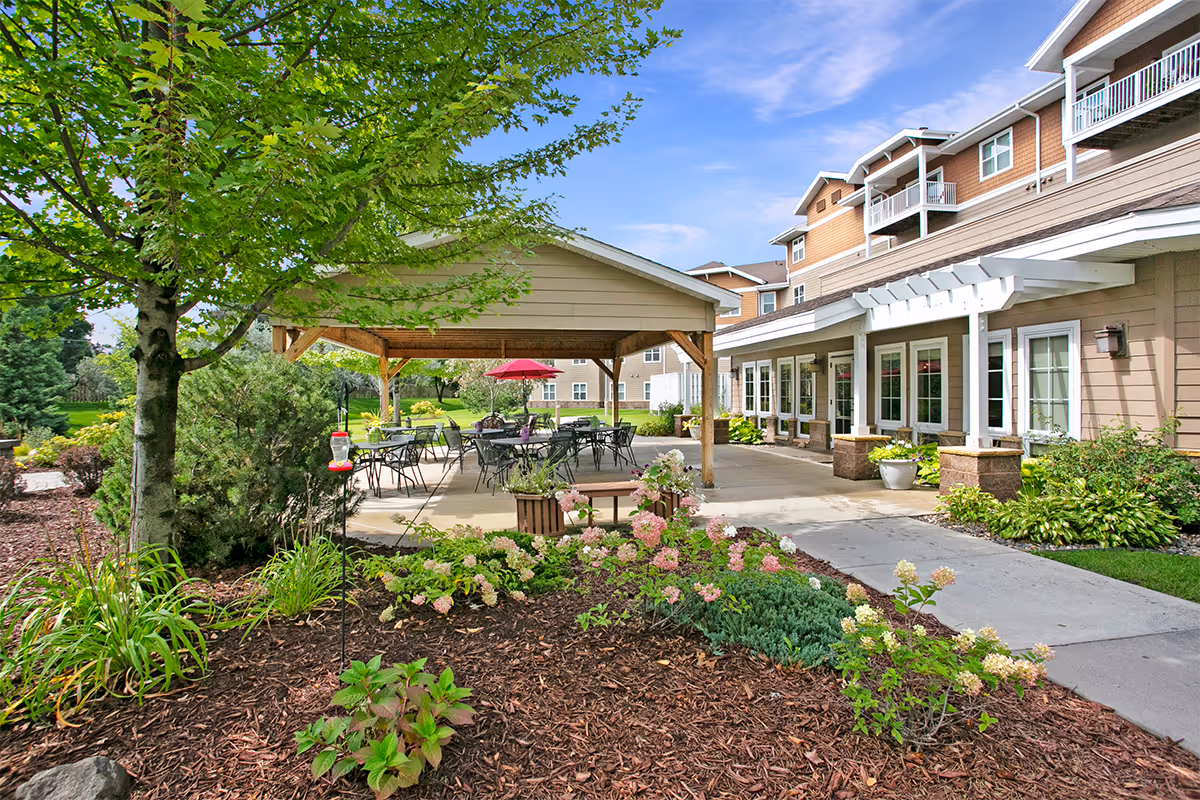 Outdoor patio area at Norris Square I facility with a covered seating area featuring tables and chairs, surrounded by landscaped garden beds with flowers and shrubs, and a multi-story building in the background under a blue sky.