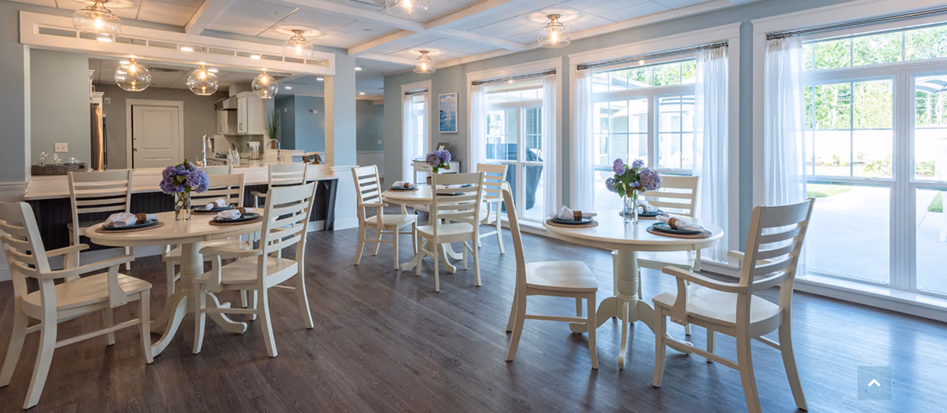 Bright and airy dining area with round tables and white wooden chairs, each table decorated with a small vase of purple flowers and place settings. Large windows with white curtains allow natural light to fill the room, and a kitchen area with a counter and bar stools is visible in the background.