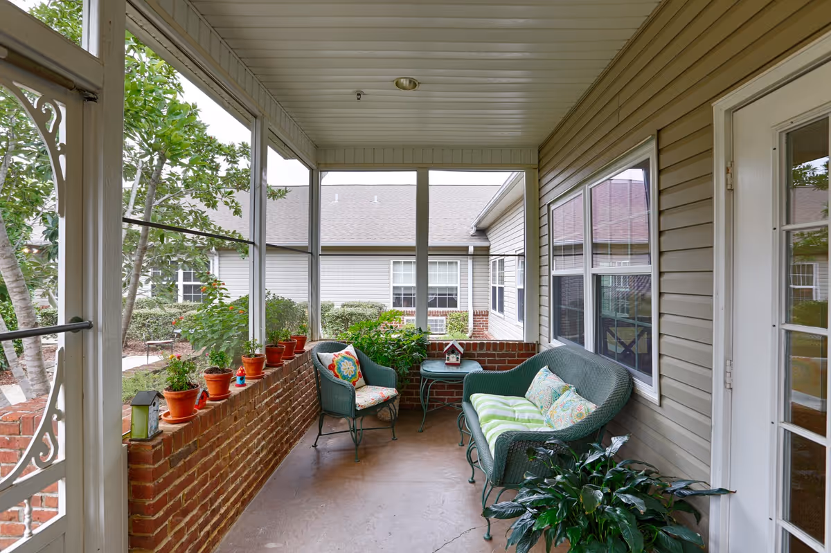 A screened-in porch area with green wicker furniture including a loveseat and a chair with colorful cushions. There is a small round table with a decorative birdhouse on it. Several potted plants line the brick half-wall, and additional greenery is visible outside the screened windows. The porch has a white ceiling and beige siding on the walls.