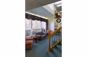 Interior view of a senior living facility lounge area with large windows letting in natural light. The room features a blue carpet, a wooden staircase with a railing, two pink upholstered chairs, a dark wooden coffee table, and a clock on the wall above the staircase landing.