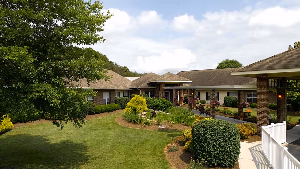 Exterior view of Knoxville Landing - A Willow Ridge Senior Living community showing a single-story brick building with a covered entrance, surrounded by well-maintained landscaping including green bushes, trees, and flower beds under a partly cloudy sky.