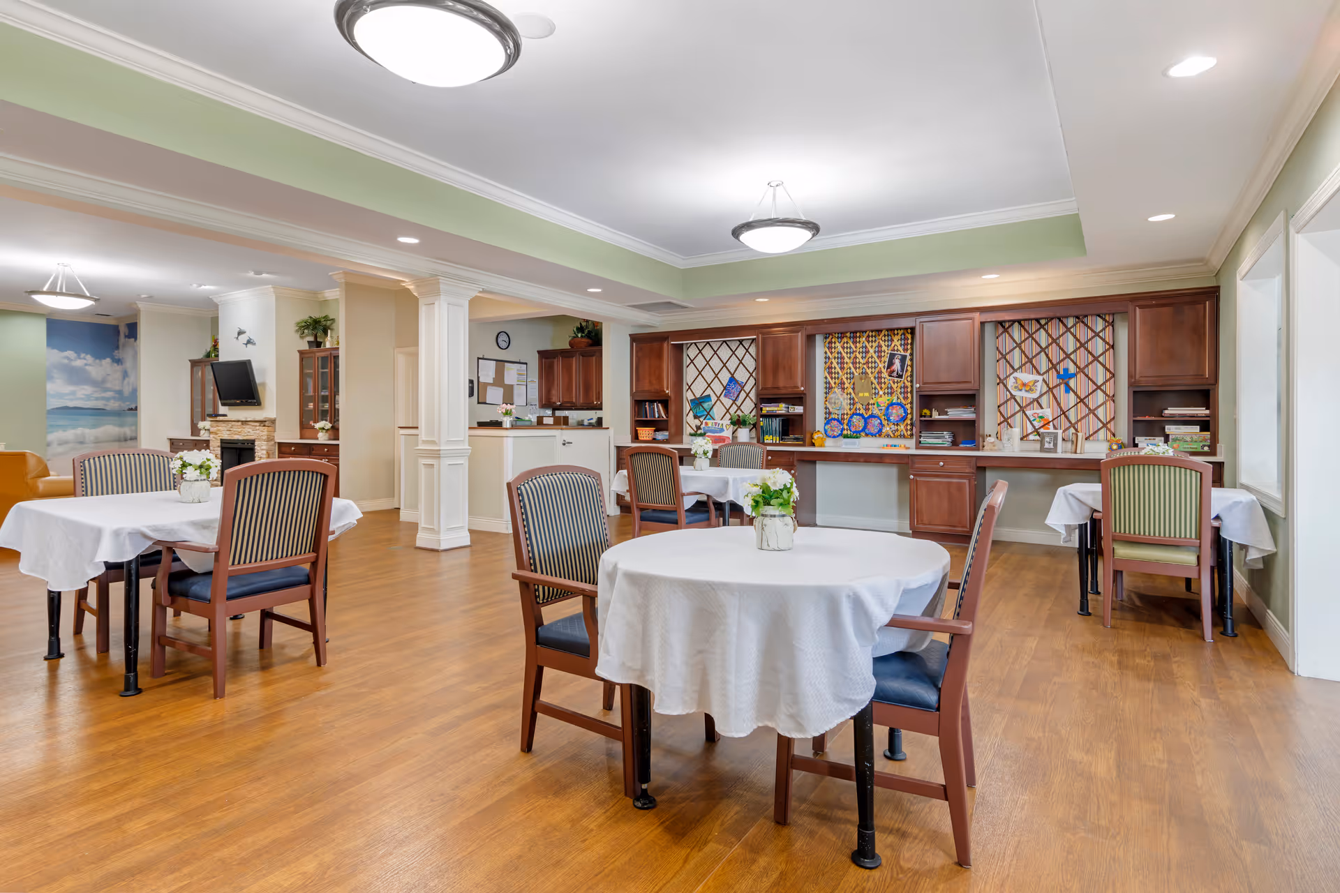 A bright and spacious common area in a senior living facility with several tables covered with white tablecloths and surrounded by wooden chairs with striped cushions. The room features wooden flooring, soft green and beige walls, ceiling lights, and built-in wooden cabinets with decorative items and artwork. A flat-screen TV is mounted above a fireplace in the background.