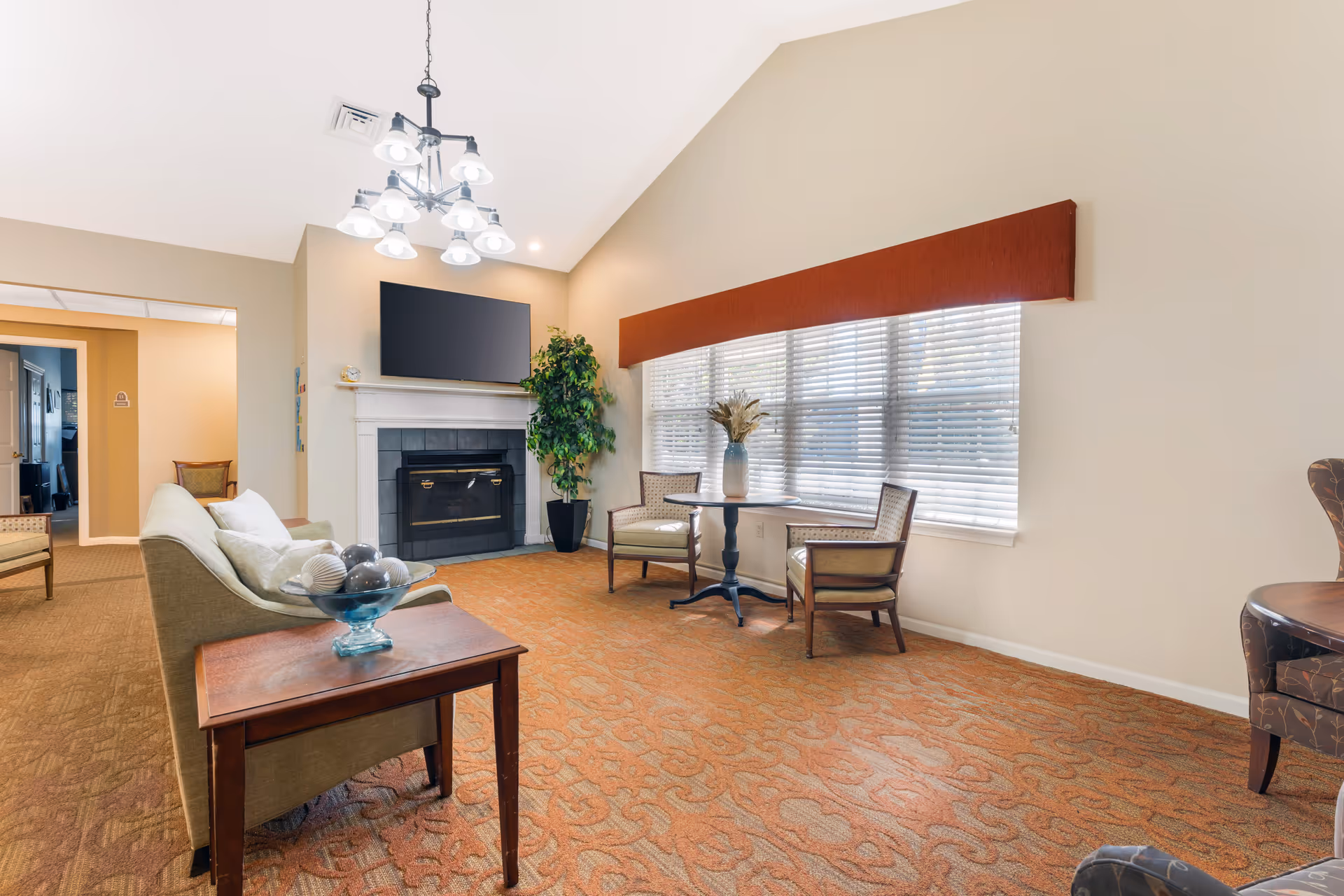 Sunlit lounge area with sofa, chairs, fireplace, wall-mounted TV, and a large window with blinds.