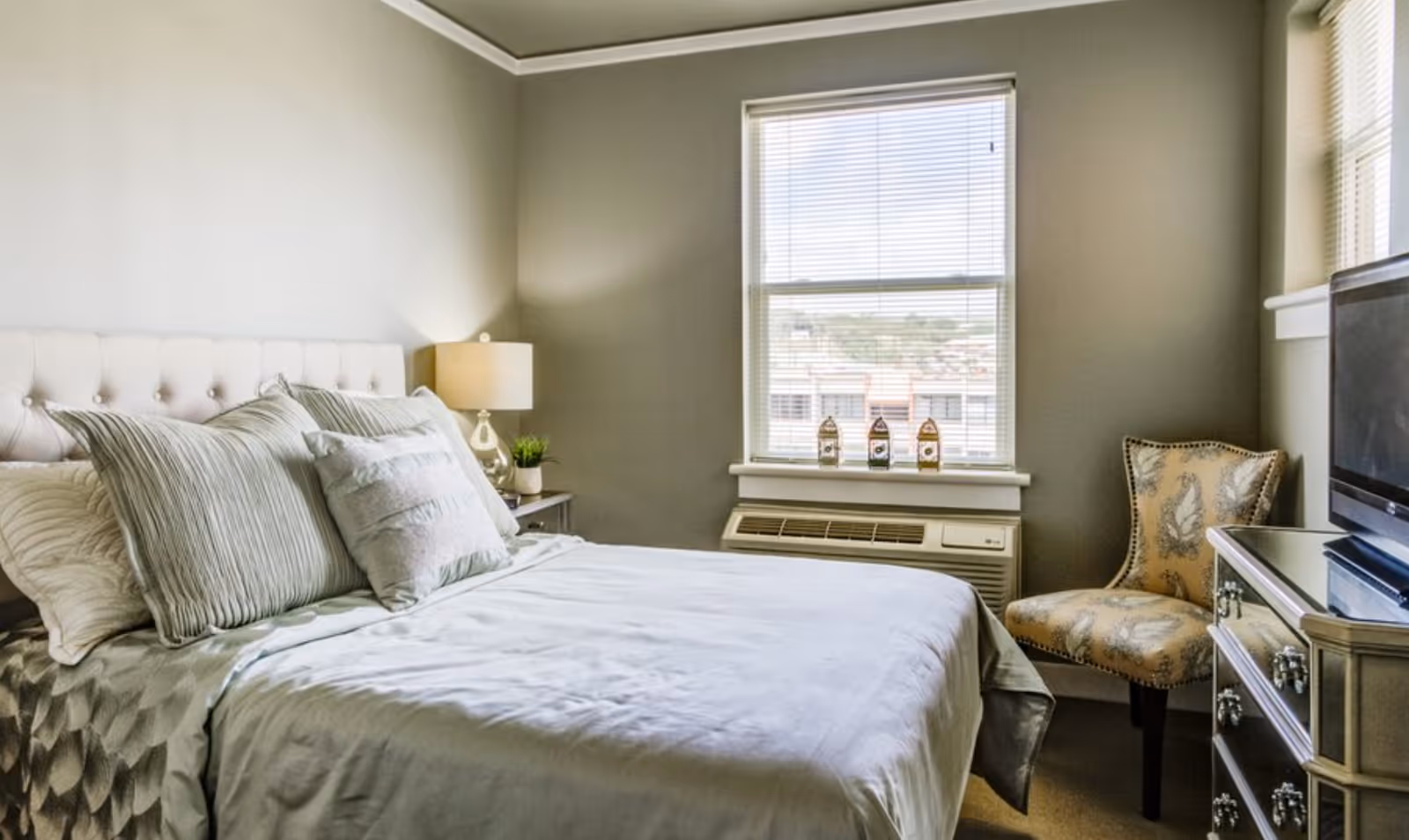 A neatly made bed with multiple pillows in a bedroom featuring a window with blinds, a bedside table with a lamp and small plant, an upholstered chair with a patterned fabric, and a mirrored dresser with a television on top.