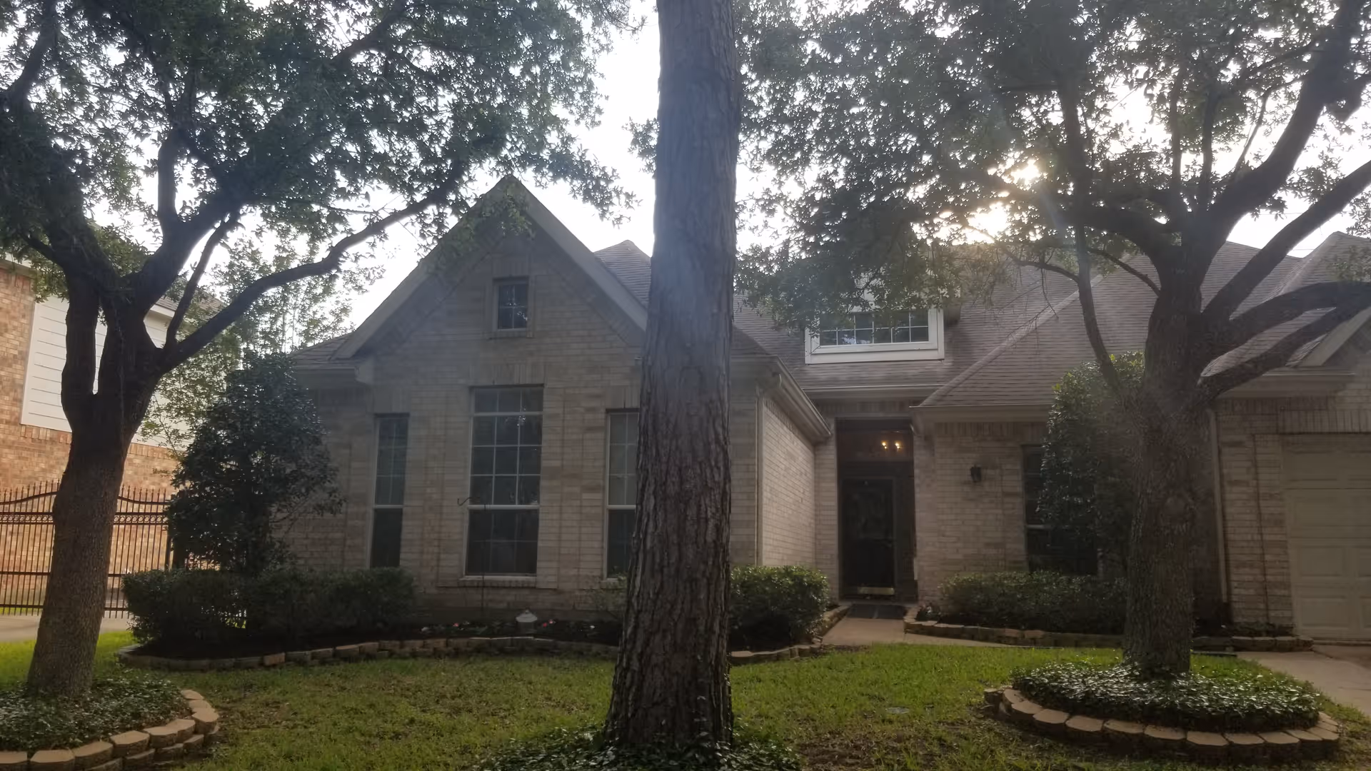 Front exterior view of a single-story brick house with a gabled roof, large windows, and a front door. The house is surrounded by a well-maintained lawn with two large trees and shrubs near the foundation.