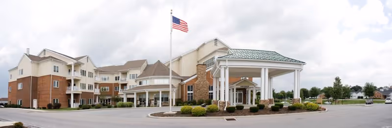 Wide exterior view of a multi-story senior living facility with a covered entrance, an American flag on a flagpole, and well-maintained landscaping under a cloudy sky.