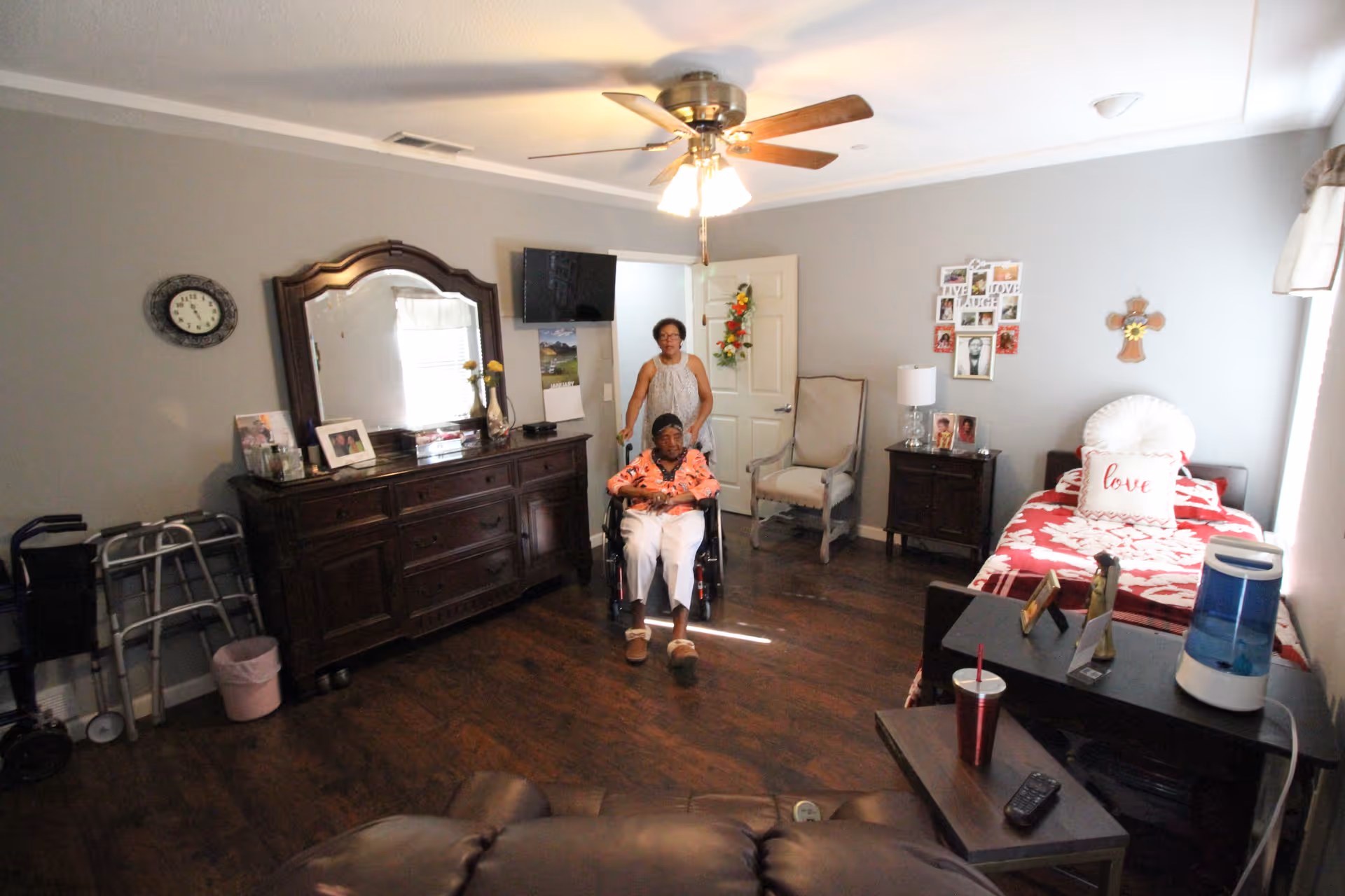 A senior living facility bedroom with two women, one sitting in a wheelchair and the other standing behind her. The room has a single bed with red and white bedding, a wooden dresser with a mirror, a wall clock, a chair, a nightstand with a lamp, and a ceiling fan with lights. There are personal items and decorations on the walls and furniture.