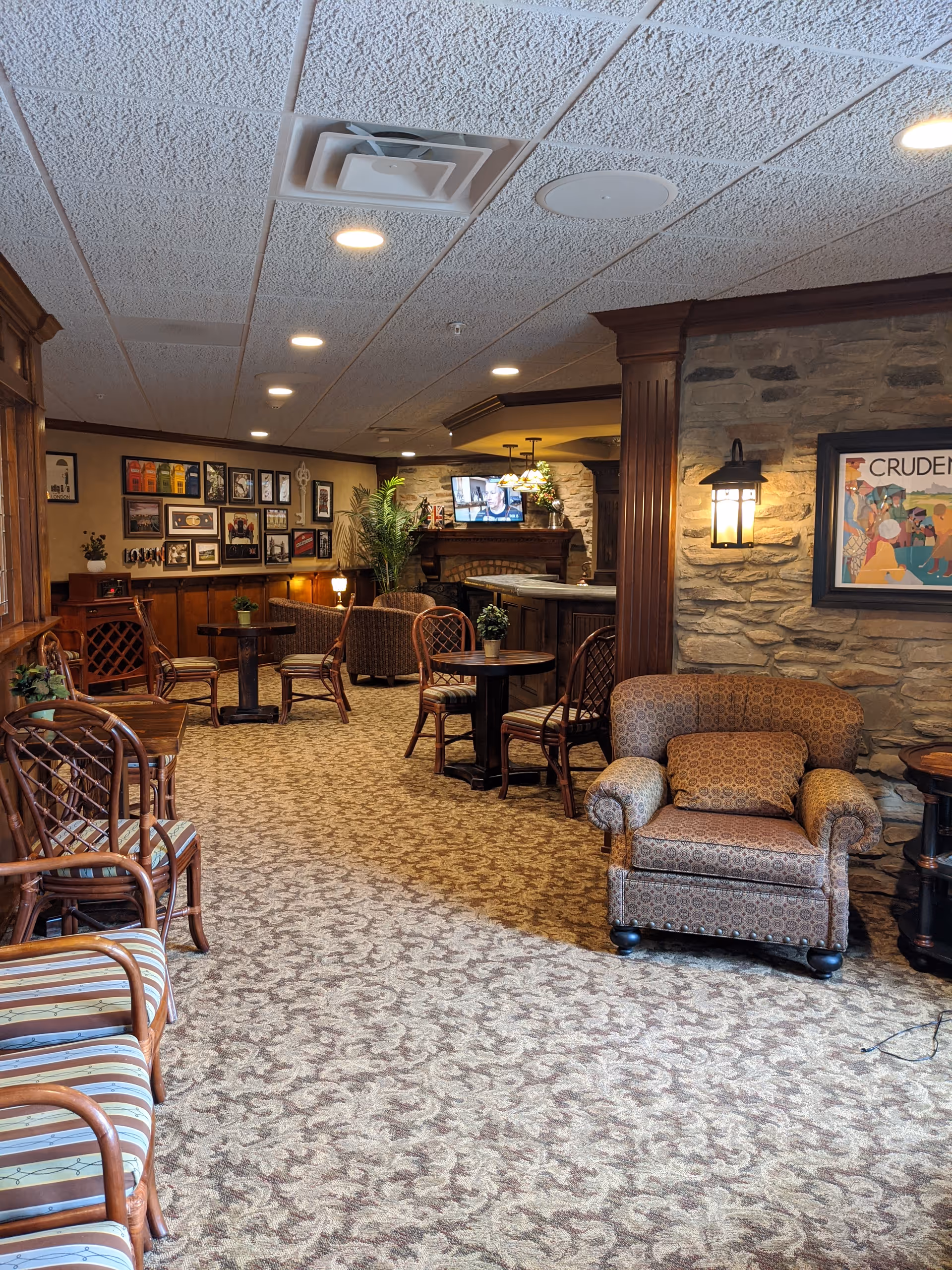 A cozy common area in a senior living facility featuring patterned carpet, wooden chairs with cushions, small round tables with potted plants, a cushioned armchair, a stone wall with a framed artwork, and a fireplace with a TV above it. The room is warmly lit with ceiling lights and wall sconces, and decorated with framed pictures on the far wall and a large plant near the fireplace.