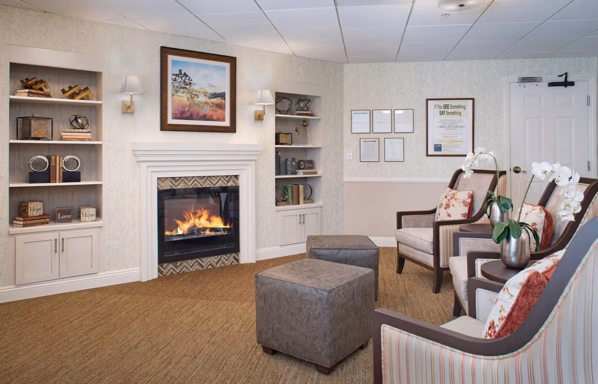 Cozy common room with a lit fireplace, built-in shelves, armchairs, ottomans and floral accents.