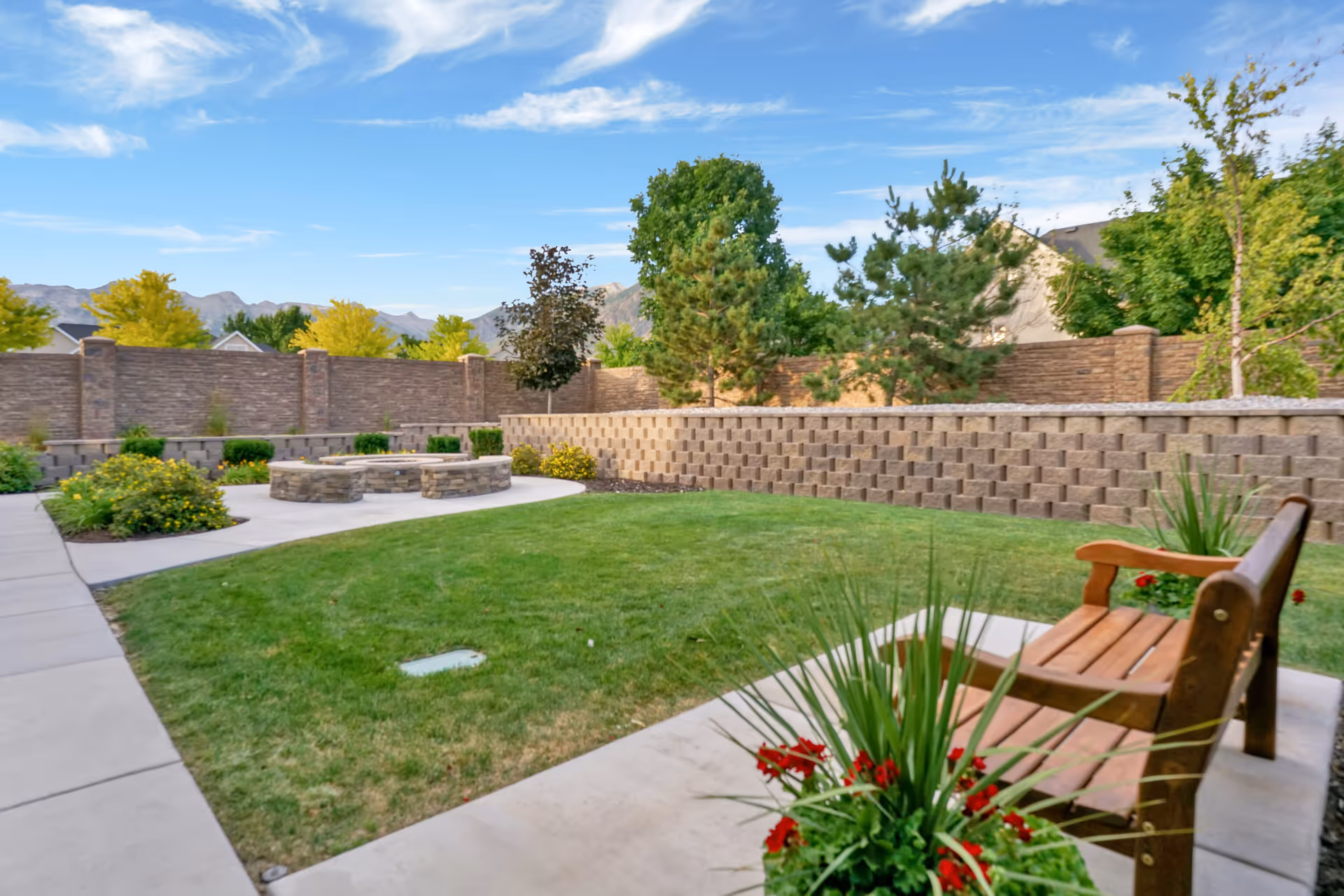 Outdoor garden area with a green lawn, a wooden bench, potted plants with red flowers, a stone fire pit surrounded by a paved walkway, and a brick wall enclosing the space with trees and shrubs along the perimeter under a blue sky with some clouds.