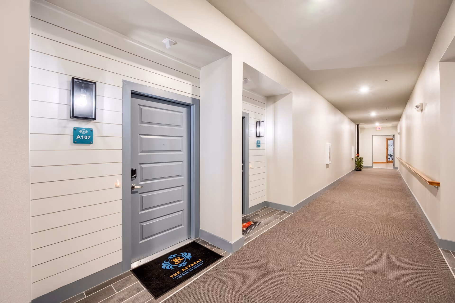 A well-lit indoor hallway in a senior living facility with a gray door labeled A107 on the left side. The door has a black doormat with a logo and text. The hallway has beige walls, a carpeted floor, a handrail on the right wall, and ceiling lights. There is a plant near the end of the hallway and an exit door in the distance.