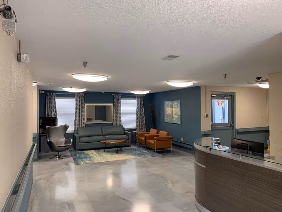 A nursing and rehab facility common area with a modern reception desk on the right, a seating area with a green sofa, two orange armchairs, a black swivel chair, a wooden coffee table, and a blue patterned rug. The walls are painted blue and beige, with two windows covered by patterned curtains and a colorful painting on the wall. The floor is a light gray marble-like finish, and ceiling lights illuminate the space.