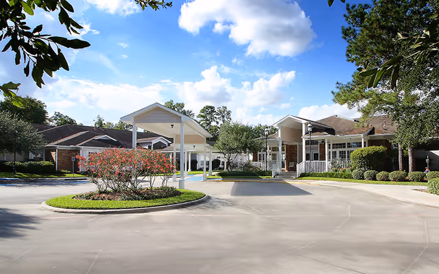 Exterior view of King's Preserve at Kingwood showing a driveway with a roundabout featuring a landscaped island with flowering bushes, surrounded by single-story buildings with covered entrances, trees, and a partly cloudy sky.