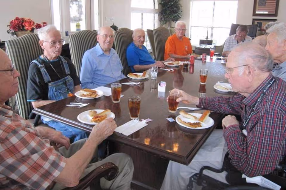 A group of elderly men sitting around a large wooden dining table enjoying a meal together. Each person has a plate with food and a glass of iced tea. The room is well-lit with large windows and decorated with flowers and framed pictures.