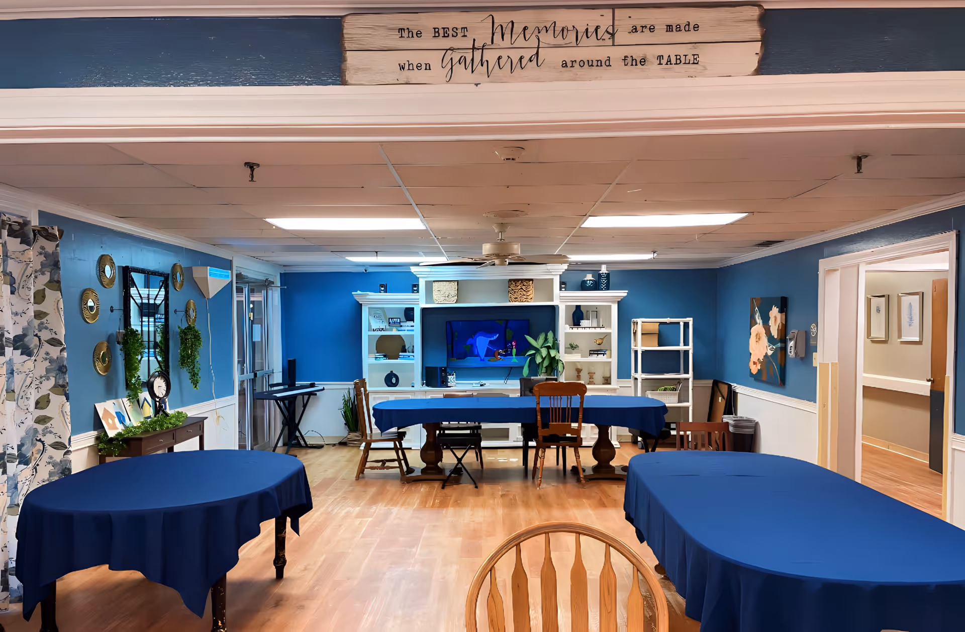A communal room with blue walls and wooden flooring featuring three tables covered with blue tablecloths and several wooden chairs. The back wall has a white entertainment center with a TV, plants, and decorative items. A sign above the room reads 'The BEST Memories are made when gathered around the TABLE'. The room is well-lit with ceiling lights and has decorative wall art and plants.