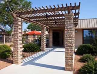Outdoor entrance area of a building with a pergola made of brick columns and wooden beams, surrounded by bushes and trees, with a clear blue sky above.
