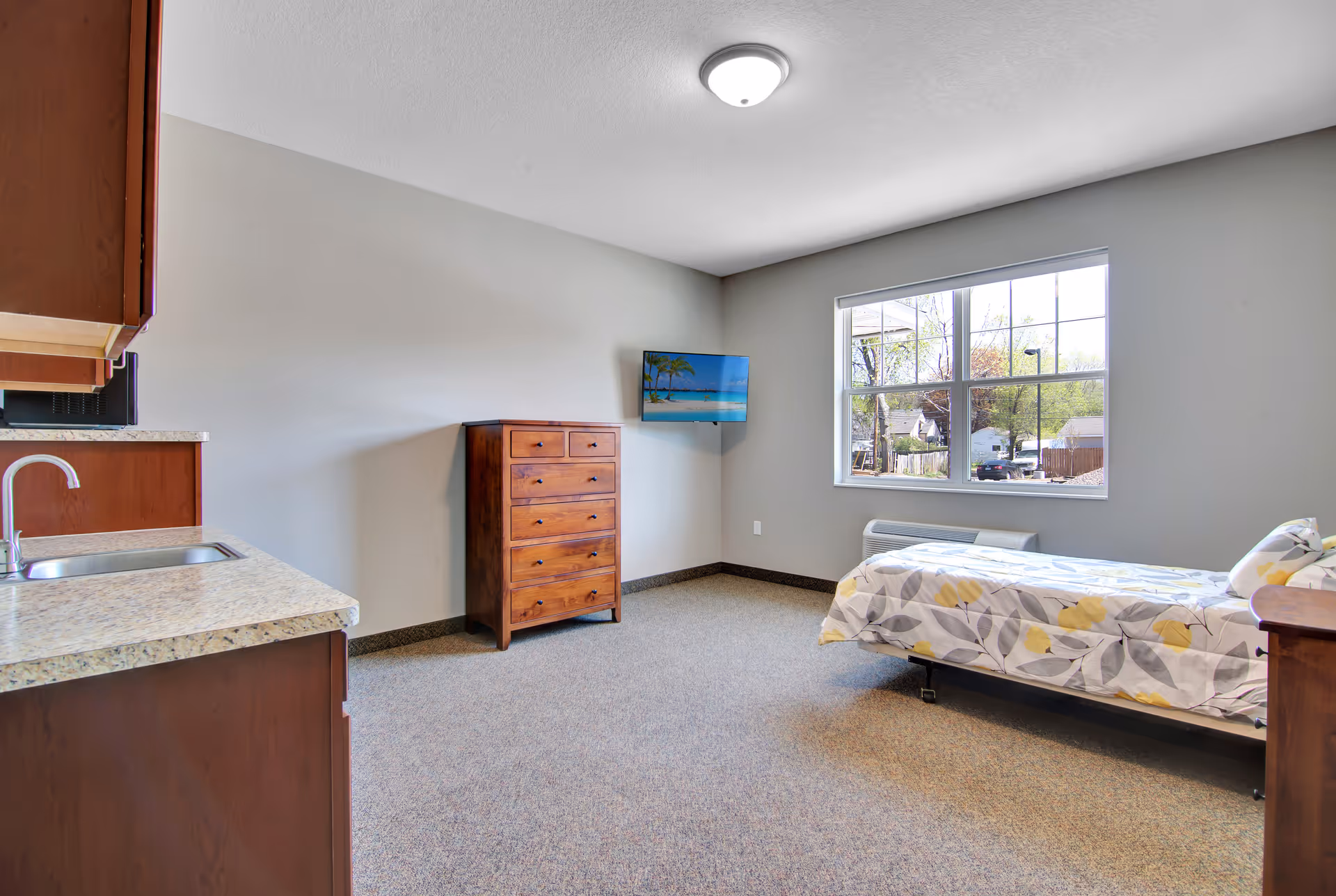 A senior living facility room with a single bed covered in a floral patterned bedspread, a wooden chest of drawers, a wall-mounted flat-screen TV displaying a beach scene, a window showing an outdoor view, and a kitchenette area with a sink and countertop.
