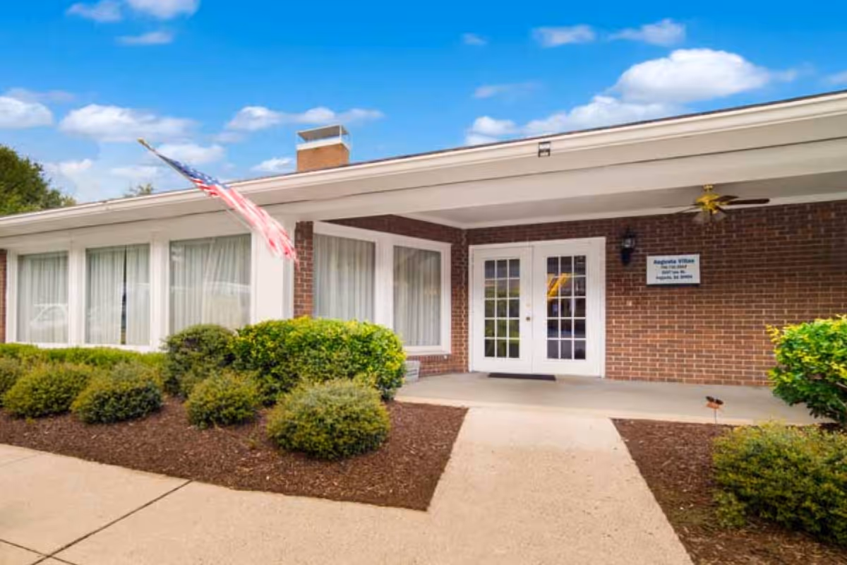 Front entrance of a single-story brick building with double glass doors, an American flag, and landscaped shrubs.