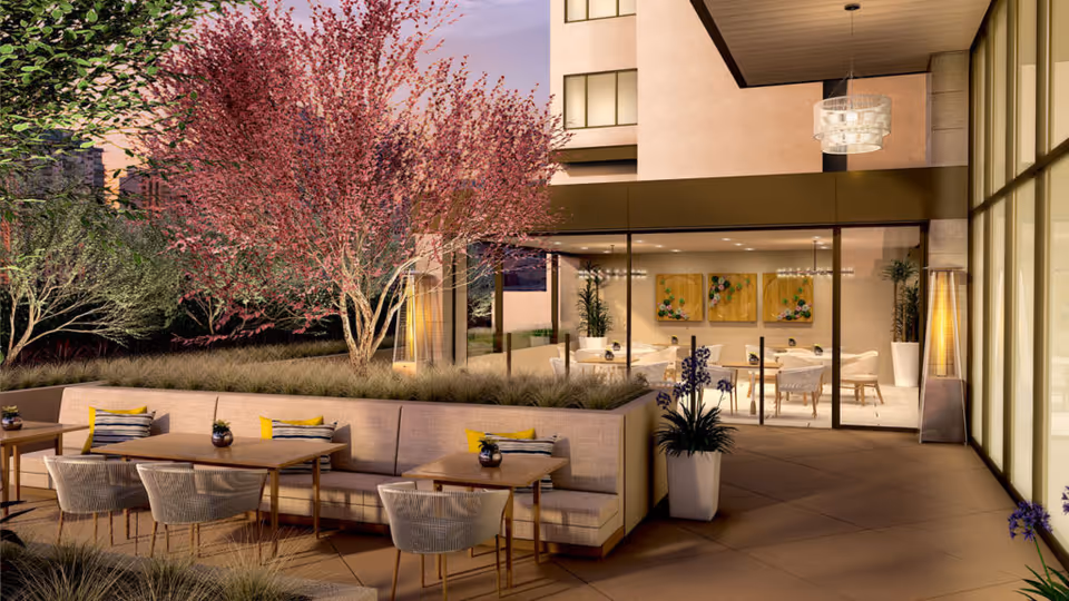 Outdoor patio with wooden tables, woven chairs, cushioned bench seating, and potted plants in front of a glass-walled dining room.