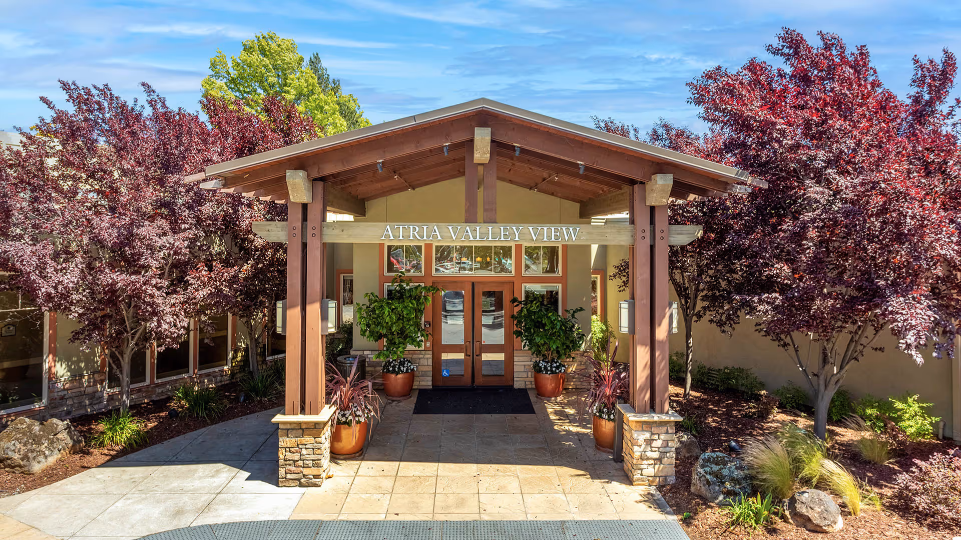 Entrance of Atria Valley View facility with a covered wooden porch supported by stone pillars, surrounded by landscaped plants and trees with purple and green foliage under a blue sky.