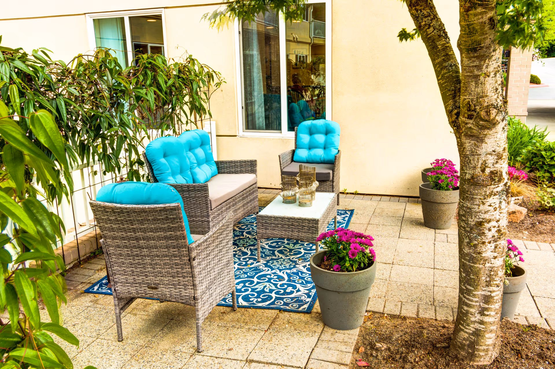 Outdoor patio area with wicker furniture including a loveseat and two chairs with bright turquoise cushions, a glass-top coffee table on a blue patterned rug, surrounded by potted purple flowers and greenery, next to a building with windows.